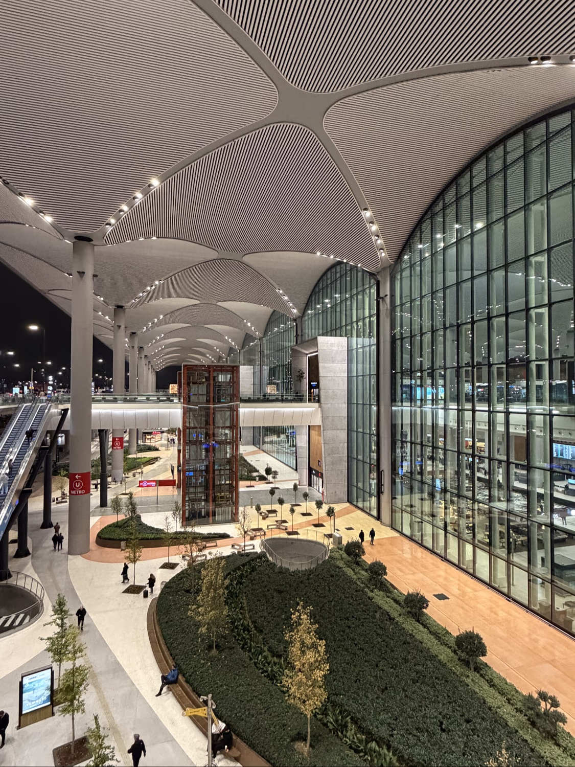 High-angle view of Istanbul Airport's vast, multi-level terminal at night, with indoor gardens and a large glass facade.