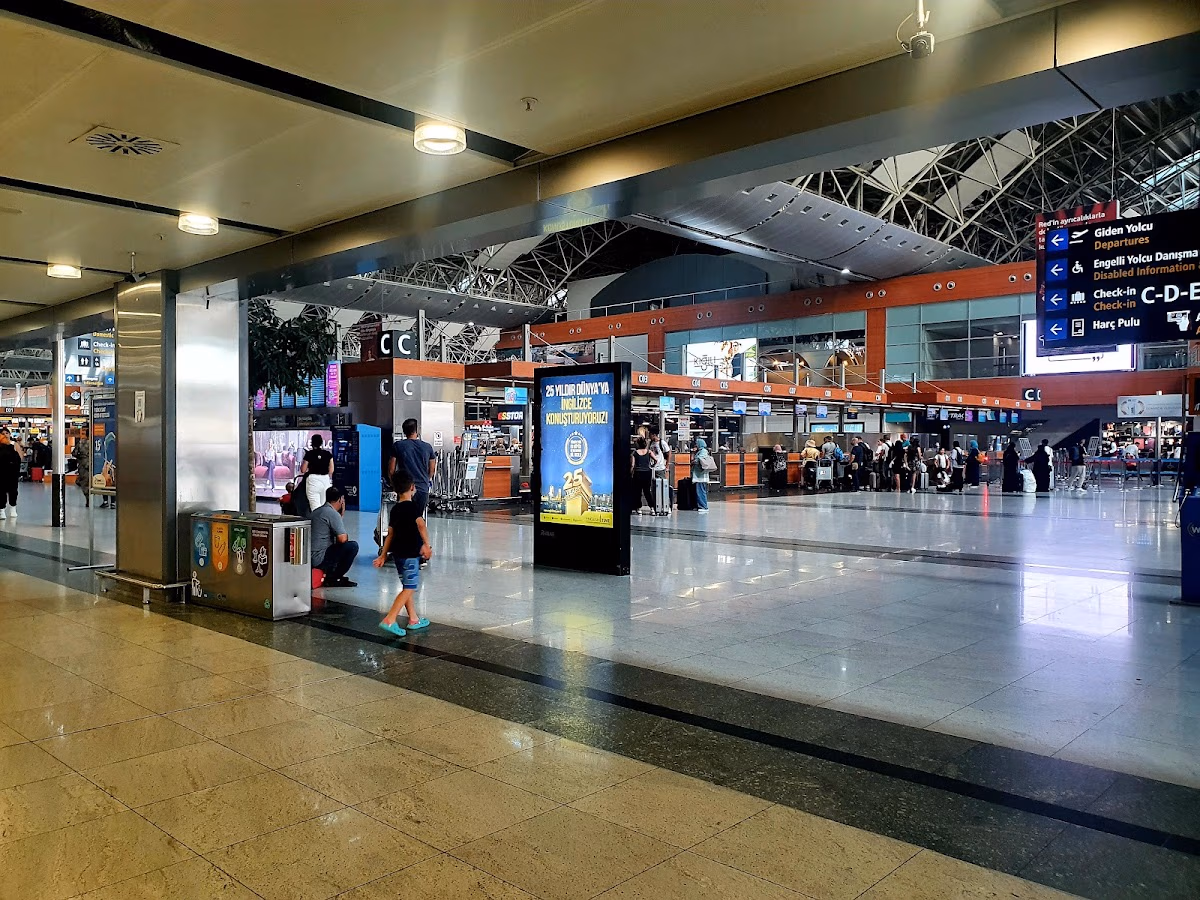 İstanbul Sabiha Gökçen Havalimanı'nın geniş check-in salonunda kontuarların ve uçuş bilgi ekranlarının önündeki yolcular.