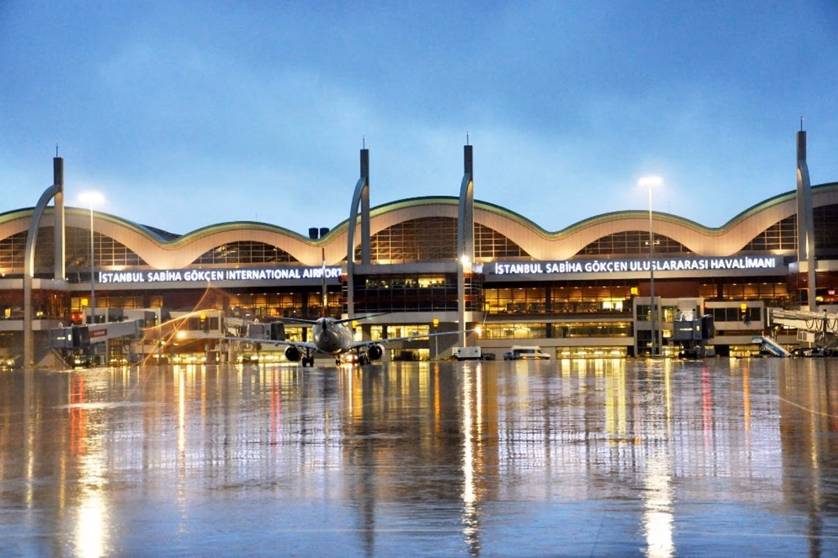 Terminal zračne luke Sabiha Gökçen osvijetljen noću, s zrakoplovom na izlazu i odrazima na mokroj pisti.