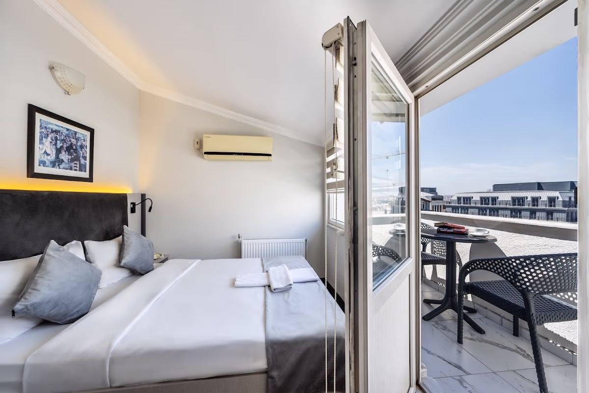 Taksim Trust Hotel'de çift kişilik yataklı, gri yastıklı, güneşli balkona açılan kapılı aydınlık ve modern otel odası.